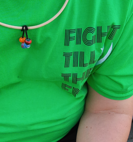Der Bildausschnitt zeigt ein grünes T-Shirt mit Fragmenten der Aufschrift "Fight till the end". Links oben ein Rattanreifen mit vier daran befestigten Glasperlen in bunten Farben.