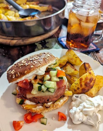 Dunkler Tisch, im Vordergrund ein weißer Essteller, darauf ein Burger mit einem dunklen Burger Bun. Die Oberseite ist etwas nach hinten geschoben, so dass geröstete Frühlingszwiebeln, veganes Patty, Käse, Gurken-Tomatensalat und eine weiße Creme sichtbar sind. Neben dem Burger befinden sich goldbraun gebackene Kartoffelspalten und ein kleiner Berg Kräuterquark. Im Hintergrund sind rechts ein Glas mit  Eistee und Eiswürfeln und links eine runde, metallene Auflaufform mit gebackenen Kartoffeln und einem Servierlöffel.