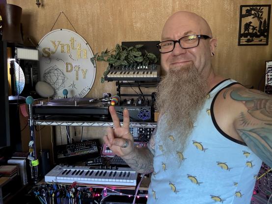 Standing in front of my turntable and small section of music gear, a large white oval sign hung on the speckled tan wall reads in gold sparkle lettering: s y n t h  d i y - I am wearing a light blue tank top patterned with tacos that have whole sharks inside them.