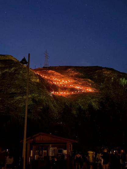 A hiking trail of people carrying the flaming wooden trunks (falles) down the mountain in the night.