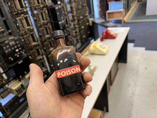 a small bottle labeled POISON held in cameraperson's hand, in front of antique telephone machinery in a museum context