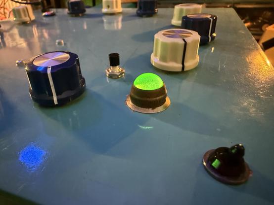 Looking across a sky blue top of an electronic instrument where large blue and white knobs can be seen next to a dome light that is currently glowing neon green.