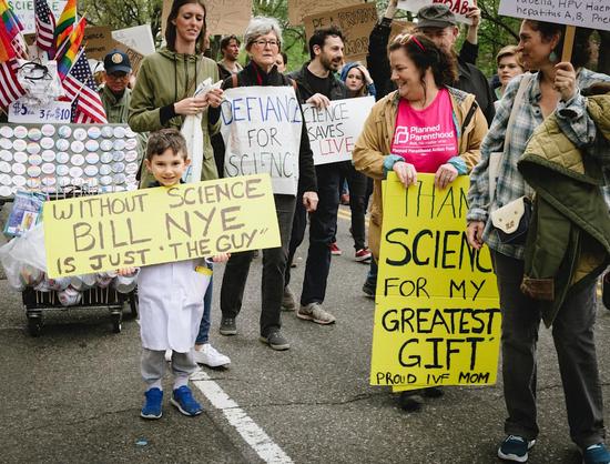 A boy holds a handmade sign saying, "Without Science, Bill Nye would just be "The Guy," while his mother's sign says, "Thank you Science for my Greatest Gift: Proud #IVF Mom."