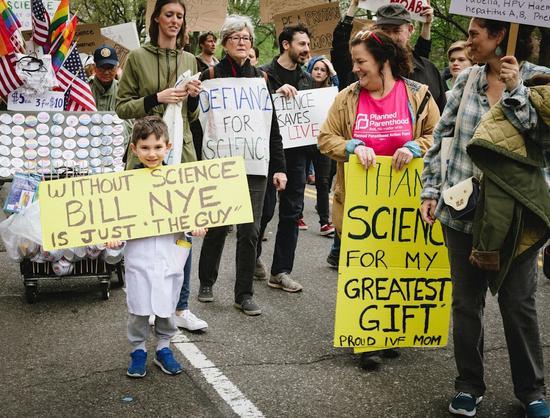 A boy holds a handmade sign saying, "Without Science, Bill Nye would just be "The Guy," while his mother's sign says, "Thank you Science for my Greatest Gift: Proud #IVF Mom."