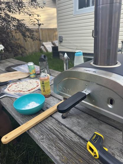 Picnic table with a pizza ready to go in the Ooni Fyra oven, a couple of craft beers also on the table.