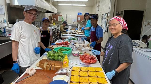 Volunteers of various ages, genders, and ethnicities are assembling sandwiches in the kitchen of the Midwest Buddhist Temple. Photo is from the temple's Facebook page.