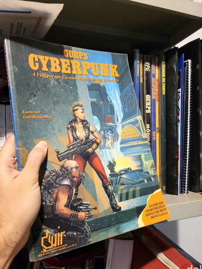 Segurado por uma mão, livro com capa ilustrada colorida em que se lê GURPS Cyberpunk: a vida por um fio num mundo de alta tecnologia. Escrito por Lloyd Blankenship. O livro que foi retido pelo Serviço Secreto dos EUA. Devir Livraria. Publicado sob autorização Steve Jackson Games.

Em segundo plano, está uma estante com mais livros relacionados a GURPS, tradicional sistema de RPG.