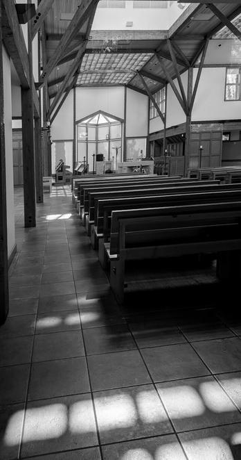 The inside of the nave of an abbey. Thin wood beams line the high ceiling and frame walls. Wood paneling and white walls come together around wood pews. Lines of rectangular light from a side window fall on the tiled floor.