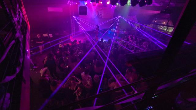 A pink-lit dance floor and stage, packed with women and nonbinary folx, lasers light up the air as performers dance on stage.