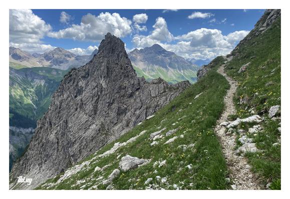 A narrow path on a steep mountain slope is leading into the void/sky. Below and next to the path is a large tooth-like rock cliff (unnamed summit). In the background the grandiose peaks on the eastern side of the Rappenalptal with green steep slopes further down. Semi-cloudy blue sky.