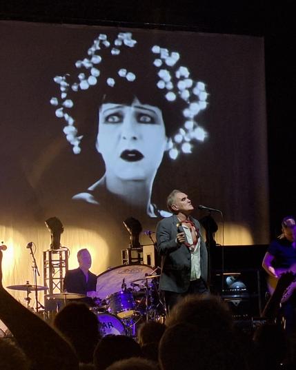 Morrissey auf der Bühne im Haus Auensee, ein Tamburin in der Hand. Im Hintergrund die Band und die schwarz-weiß Videoprojektion einer irritiert blickenden Frau