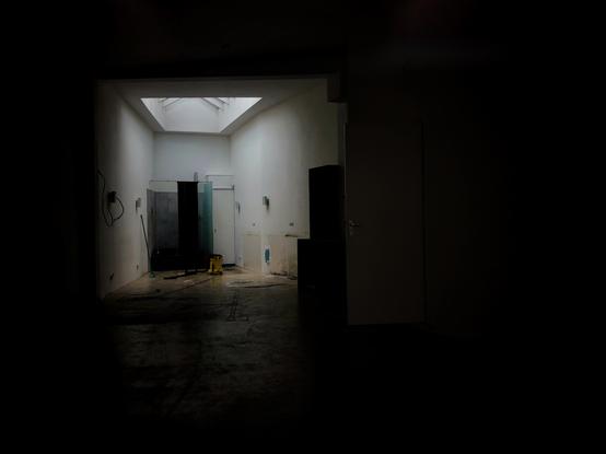 Dark image of a small, empty shop interior. At the far end of the room an area is light up by a skylight above. Visible in this area are what's left of a shower stall with glass and grey tiles on the wall. A little yellow cleaning machine can also be seen in front of the shower.