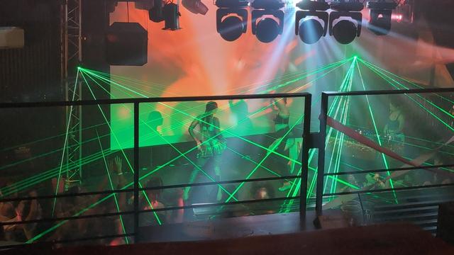 A green and orange-lit dance floor and stage, packed with women and nonbinary folx, lasers light up the air as performers dance on stage.