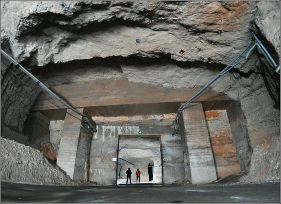 Deux silhouettes humaines se tiennent debout sous une arche de béton, soutenant le ciel d'une galerie en carrière de craie.