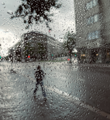 A rainy city street scene viewed through a glass wet with water droplets and obscuring the view. A person walks in the background.
