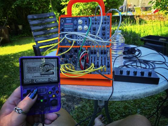 A sun-drenched backyard scene featuring a bright orange Eurorack modular synth case on a round white garden table. The synth is intricately patched with yellow cables for CV, gray ones for audio, and a green cable linking the internal mixer to an FX unit. To the right sits a compact stereo mixer where the drone signal and the R36s handheld console come together as audio sources. A full water bottle glistens beside the gear, catching the light. In the foreground, a hand with shimmering deep blue gel nails and silver rings holds a translucent purple R36s handheld console, its screen proudly showing the title screen of "Super Mario Land 2: 6 Golden Coins". At the bottom, bare legs peek into the frame—knees bent, toes relaxed on the grass. The whole setup oozes chaotic creative energy, blending tactile audio wizardry with pixelated retro gaming under the open sky.