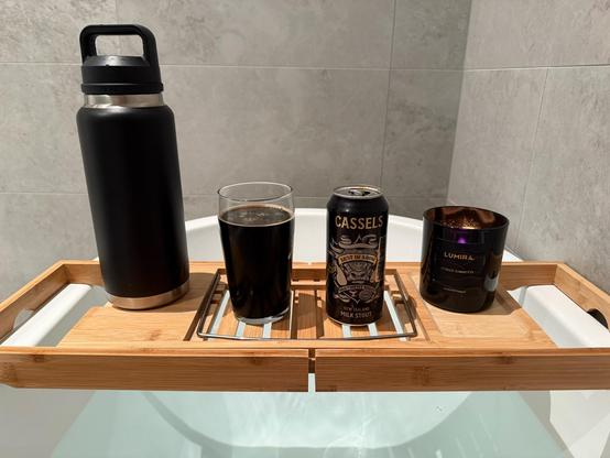 An image showing a range of items on a bath table. On the left there is a large black yeti water bottle. Next to that is a large pint glass filled with a dark thick beer. Next to that is the cassels milk stout can and on the right is a “Cuban tabacco” candle which is lit.
