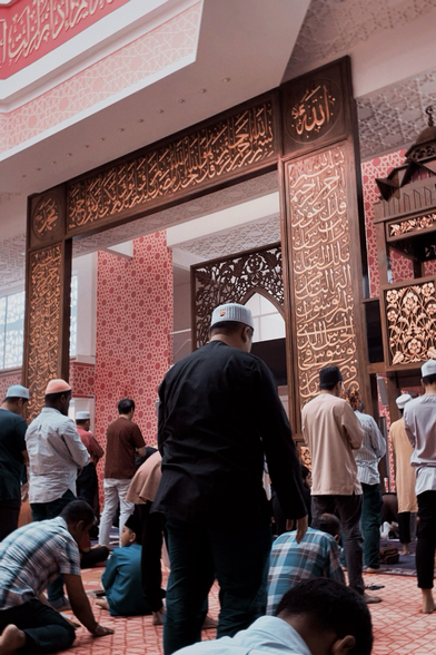 The image depicts the interior of a mosque with a group of people engaged in prayer. The central focus is on a large wooden archway with intricate Arabic calligraphy, featuring the name of Allah and other religious inscriptions. The walls are adorned with red and white geometric patterns, and the ceiling has a decorative design. The floor is covered with a red patterned carpet. People are seen in various positions of prayer, with some standing and others kneeling or sitting on the floor. The individuals are dressed in traditional attire, including caps and long garments, with some wearing masks. The overall atmosphere is one of reverence and devotion. Provided by @altbot, generated privately and locally using Ovis2-8B 🌱 Energy used: 0.176 Wh