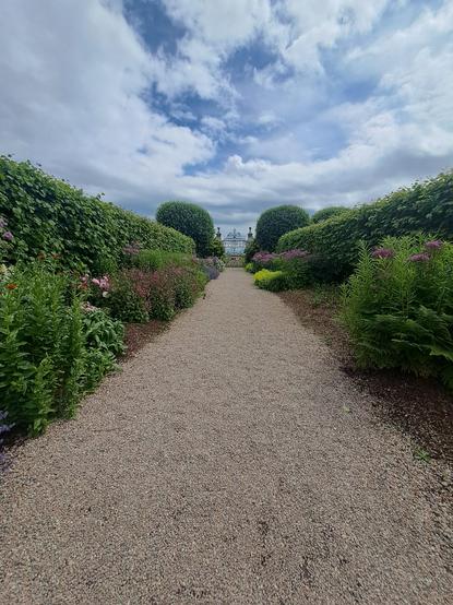 A reddish brown gravel pathway leads the viewer to a decorative iron gate in the middle distance. Green hedges line the pathway on both sides, while on the inner side of the hedges are some beautiful flowers, mostly purple in colour, with a couple of pinks and a white flower bush towards the end of the path. Larger, round green shrubs cut into a round shape are growing at the end of the hedges, just before the gate, partially blocking the grey stone pillars that are sitting either side of the gate. The sky overhead is mostly covered in greyish white clouds, with just a bit of blue peeking through. 
During the recent trip to Scotland with my wife we visited the lovely Dunrobin Castle on our way from Wick to where we where staying near Invergordon. The gardens were a big draw card for us, and they are, indeed, very lovely.