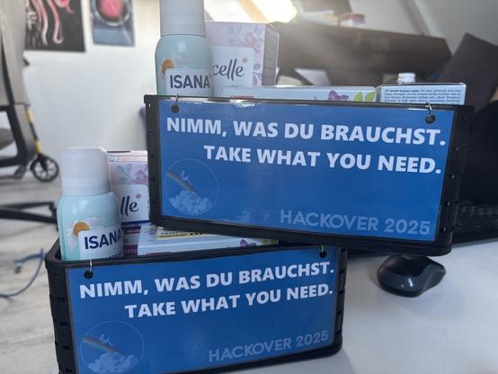 Auf einem Tisch stehen zwei kleine Kisten übereinander.
Auf der Vorderseite ist jeweils ein Schild angebracht auf dem steht: „Nimm was du brauchst. Take what you need.“
In der Box sind Hygieneartikel zu erkennen.