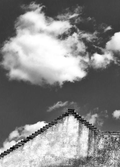 This black and white photograph captures a stark, minimalist view of a building's gabled roofline against a dramatic sky.

The prominent feature is the triangular peak of a building, seen from a low angle looking upwards. Above the roof, the sky is filled with a dynamic arrangement of clouds. There's a large, bright, billowy cloud dominating the upper left portion of the frame, and smaller, more scattered clouds elsewhere.
