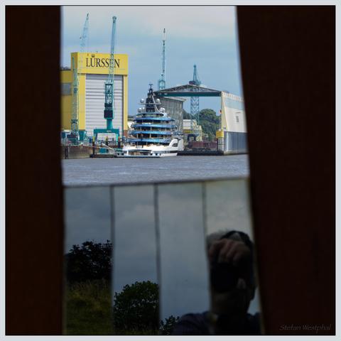 Eine luxuriöse Yacht liegt in der Nähe einer Werft mit großen Kränen vor Anker, auf denen der Name des Unternehmens Lürssen prangt. Die Szene wird teilweise von spiegelnden Flächen eingerahmt, die das umliegende Grün, den Himmel und den Fotografen einfangen.

A luxurious yacht is anchored near a shipyard with large cranes emblazoned with the name of the Lürssen company. The scene is partially framed by reflective surfaces that capture the surrounding greenery, the sky and the photographer.