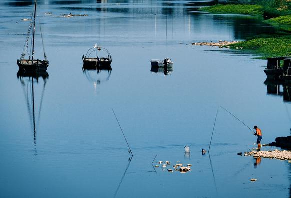 A tranquil scene of a river with several boats anchored. In the foreground, a person is fishing from the shore, while reflections of the boats and surrounding scenery can be seen on the calm water.