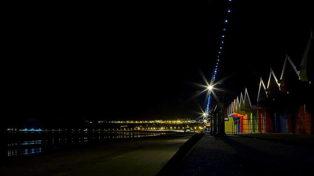 Night view of a seafront promenade with lit beach huts on the right and distant town lights reflecting across a calm bay.