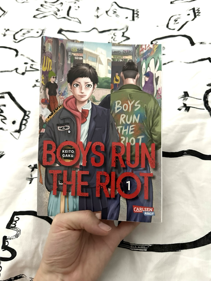 Photo of a hand holding up the first volume of the manga “Boys Run the Riot” by Keito Gaku. The cover art shows a trans boy with short hair, who wears a masculine casual outfit on one and a feminine school uniform on the other side. Behind him stands another boy with a man bun with his back to the viewer. He wears a jacket with the text “Boys Run the Riot” on it.