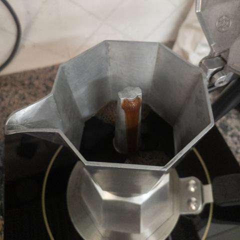 A silvery aluminium moka pot standing on an induction hot plate with a stainless steel adapter, spewing coffee from its spout on the upper chamber.
