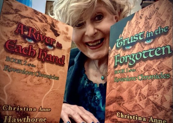 In the background is an older woman with wavy blonde hair who's smiling and is dressed blue.

In the foreground on the right is a print copy of Trust in the Forgotten, Book 1 of the Kovenlore Chronicles with the author's name below. The text is in a blue-green, the author's name in a dark yellow. The image is of a golden negative of a hand drawn map. There are jade eyes in the title.

In the foreground on the left is a print copy of A River in Each Hand, Book 2 of the Kovenlore Chronicles with the author's name below. The title is in red, the subtitle in blue-green, the author's name in a dark yellow. The image is, again, that of a golden negative of a hand drawn map. There are jade eyes in the title.