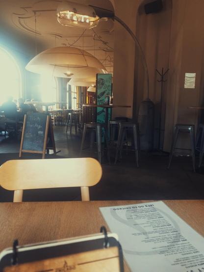 Na snímku je útulný interiér restaurace s dřevěnými stoly a židlemi, kolem kterých jsou rozestavěné kovové stoličky a stojanová tabule s nápisem „Thirsty Thursday“. Z prostoru nad stoly visí velké polokulovité stropní lampy, které společně s denním světlem pronikajícím velkými obloukovými okny vytvářejí teplou a klidnou atmosféru.