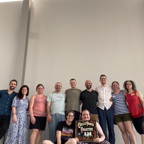 11 people smile at the camera. 9 stand in a row. Two sit in the front with a “queer tango collective” sign.