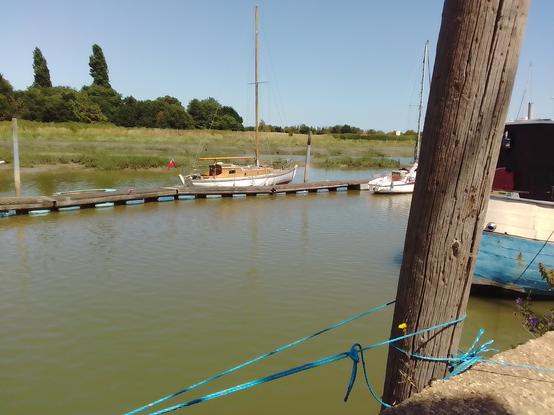 Per body, an old wooden yacht alongside a pontoon, Marsh and sea wall in the background, and our rowing tender tied up after of it.