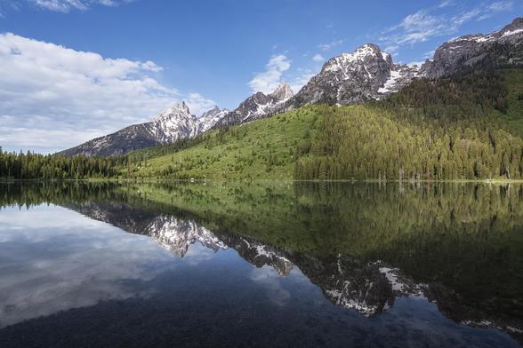 Majestic snow-capped Teton mountains rise above lush green forests and are perfectly mirrored in the calm, reflective waters of String Lake in Grand Teton National Park.