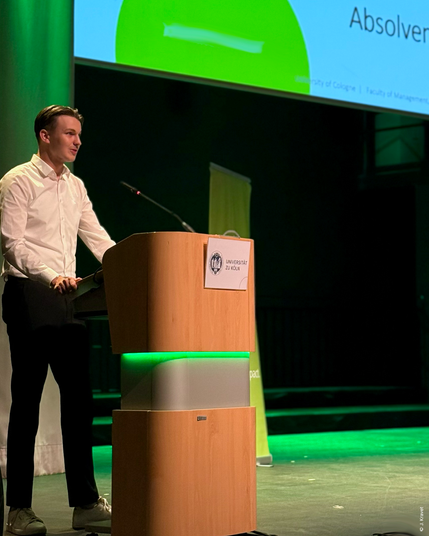 Young man in a white shirt and dark trousers speaking at a wooden podium lit by green lights, in front of a presentation slide reading “Absolvent” at the exam celebration of the Faculty of Management, Economics and Social Sciences, University of Cologne.