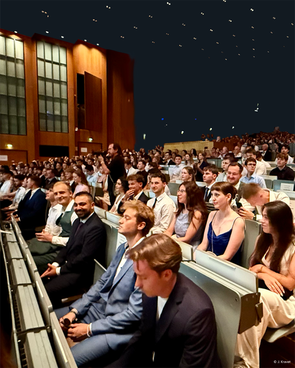 Large group of formally dressed WiSo graduates and guests seated in a modern auditorium with tall wooden walls and large windows, under a dark ceiling dotted with small star-like lights, during the exam celebration at the University of Cologne.