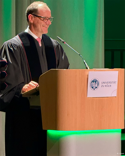 Prof. Werner Reinartz, PhD speaking at a wooden podium lit by green lights at the exam celebration of the Faculty of Management, Economics and Social Sciences, University of Cologne.