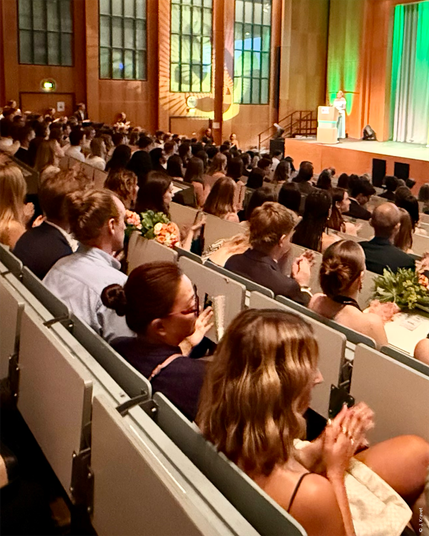 Large group of formally dressed WiSo graduates and guests seated in a modern auditorium with tall wooden walls and large windows, watching young woman in a white dress speaking at a wooden podium in the background.