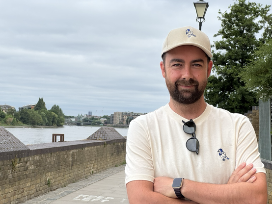 Dani standing in front of a riverside wall with his arms crossed, wearing a cream t-shirt and matching cap embroidered with a stylized blue Mandarin duck logo. A pair of sunglasses hangs from his shirt, and he’s smiling slightly at the camera. Trees, water, and the Hammersmith bridge can be seen in the background under a cloudy sky.