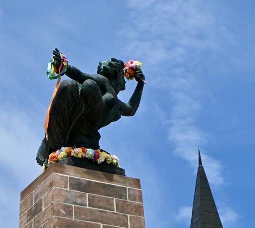 Eine Bronzestatue in einer dynamischen Pose, geschmückt mit einem bunten Blumenkranz. Sie streckt eine Hand nach hinten aus, um das Handgelenk sind bunte Bänder befestigt, während sie in der anderen Hand einen bunten Blumenstrauß hält. Die Statue befindet sich auf einem hohen Backsteinsockel. Ein nahe Turmspitze ist ebenfalls sichtbar. Der Himmel ist sommerlich blau mit weißen Wölkchen.