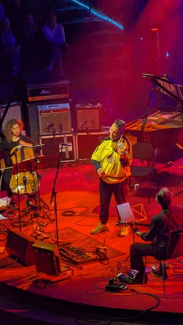 Nigel Kennedy Violine  and Beata Urbanek Kaliowska Cello on stage, which is illuminated in red light. Kennedy weares a yellow shirt, black trousers and yellow trainers.and plays the violin. Urbanek Kaloowska, dressed black sits on the left side of the stage an plays the cello. In.the background is a piano.