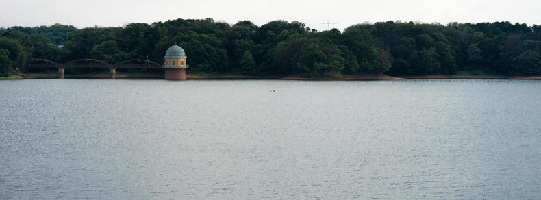 Calm body of water with a small, domed building on stilts near the shoreline, surrounded by dense, green trees. A bridge with multiple arches connects the building to the forested land. Overcast sky above, with the distant outline of hills in the background.