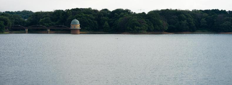 Calm body of water with a small, domed building on stilts near the shoreline, surrounded by dense, green trees. A bridge with multiple arches connects the building to the forested land. Overcast sky above, with the distant outline of hills in the background.