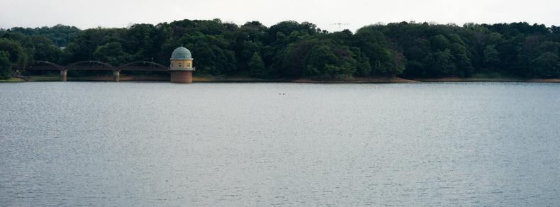 Calm body of water with a small, domed building on stilts near the shoreline, surrounded by dense, green trees. A bridge with multiple arches connects the building to the forested land. Overcast sky above, with the distant outline of hills in the background.