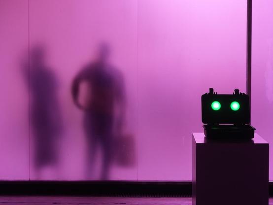 Der Grundton des Bildes ist Pink. Als Schatten sieht man zwei Personen im linken Teil des Bildes. Eine Person trägt eine Tasche. Im rechten Teil steht ein Gerät auf einem Podest mit zwei grünen Lampen.