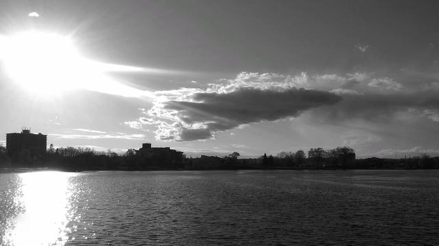 The glare of the sun is seen over a lake with a dark cloud to the right. The silhouette of buildings and trees can be seen in the background and the glare of the sun in the water. This is a black and white image.