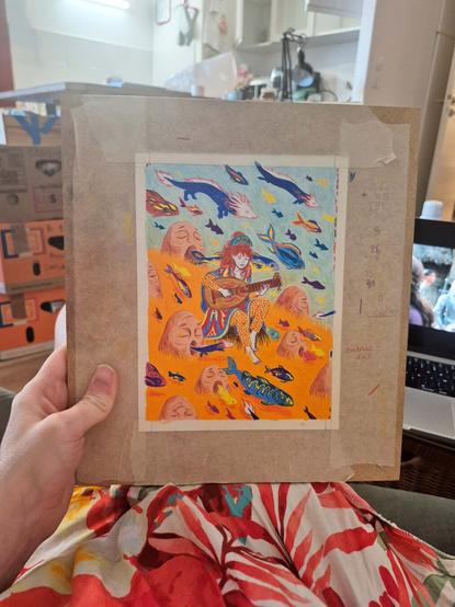 Here’s a photo of a freshly finished colored pencil drawing. It shows a character sitting on a rock, playing a lute, surrounded by colorful fish emerging from the mouths of large stone heads that appear to be singing. The drawing is still taped to its working board with masking tape, and I’m holding it on my lap, over a flowery dress.
The whole piece features a vivid palette of blues, oranges, reds, and yellows.
I kept all my little notes around the edges, as usual.