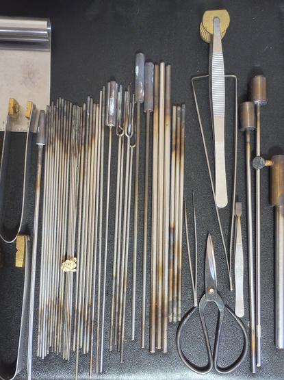 Image of a range of steel tools used in hot glass art (lampworking), set against a black tabletop background.