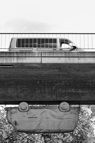 Ein Schwarz-Weiß-Foto zeigt eine Skulptur eines Kleinbus, die unter einer sehr massiven Beton-Brücke hängt. Oben auf der Brücke fährt ebenfalls ein Kleinbus in die entgegengesetzte Richtung. Davor sieht man ein Brückengeländer. Im Hintergrund ist nichts erkennbar außer Himmel und Grünpflanzen.

Die Bildkomposition ist minimalistisch und konzentriert sich auf das Auto, das unter der Brücke hängt. Der Fokus liegt auf der ungewöhnlichen Perspektive und der geometrischen Form des Bildes. Das Auto ist in der Mitte des Fotos positioniert und hängt scheinbar in der Luft, was eine unerwartete und interessante Szene erzeugt.